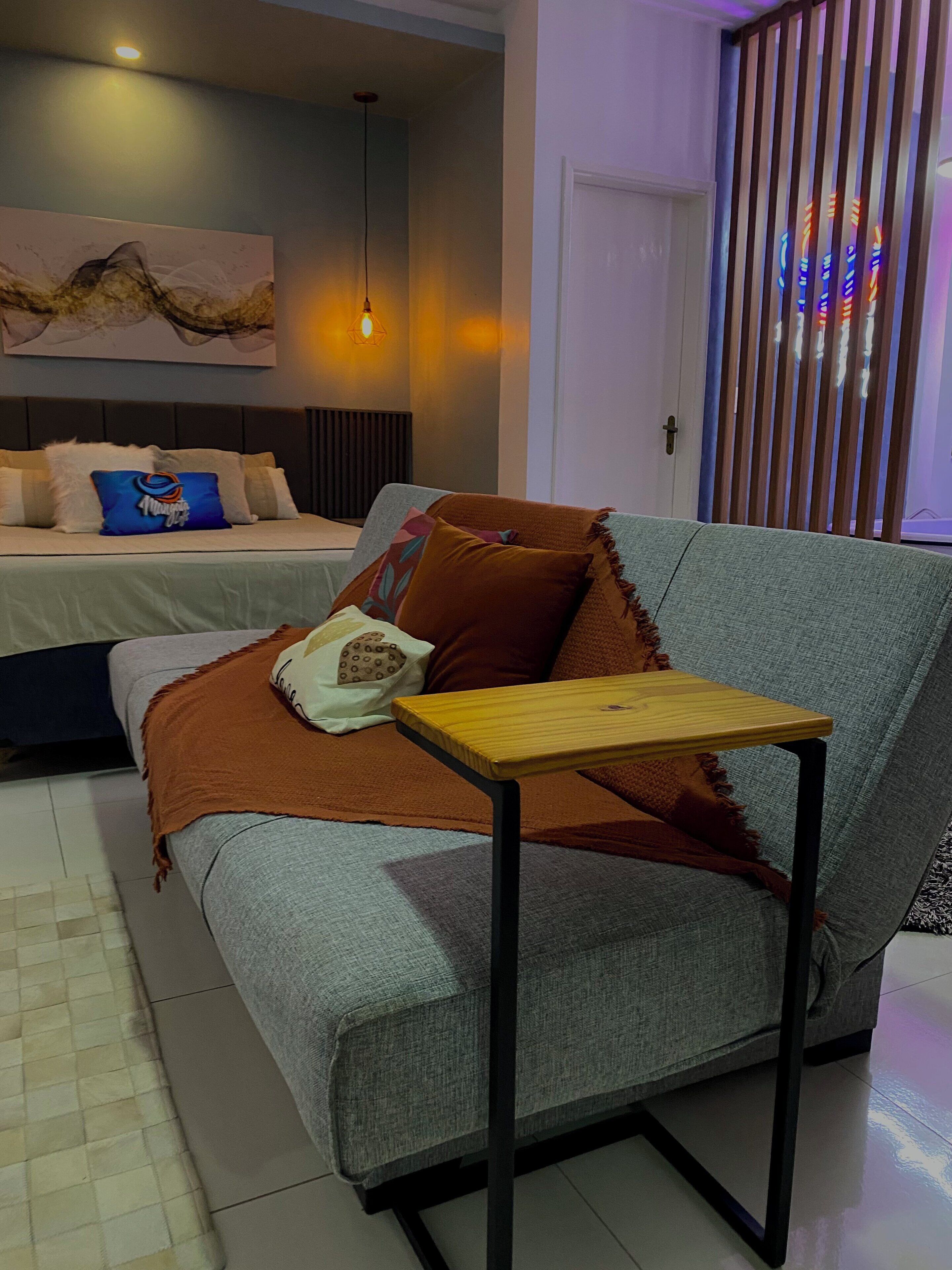 A modern bedroom with a grey sofa, a wooden side table, and a bed with multiple pillows.
