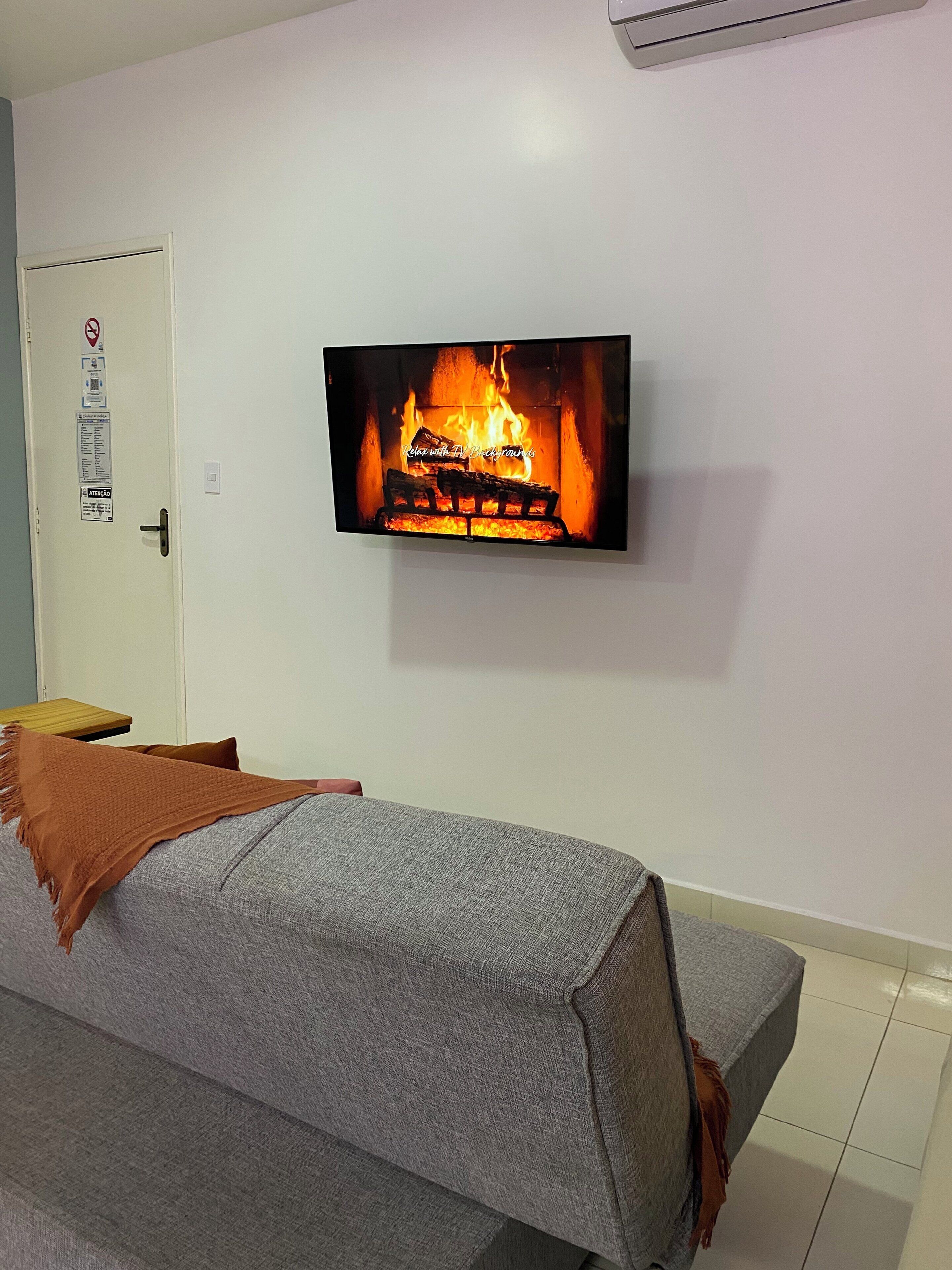 Habitación con una televisión mostrando una chimenea, un sofá con una manta y una puerta con un cartel de prohibido fumar.