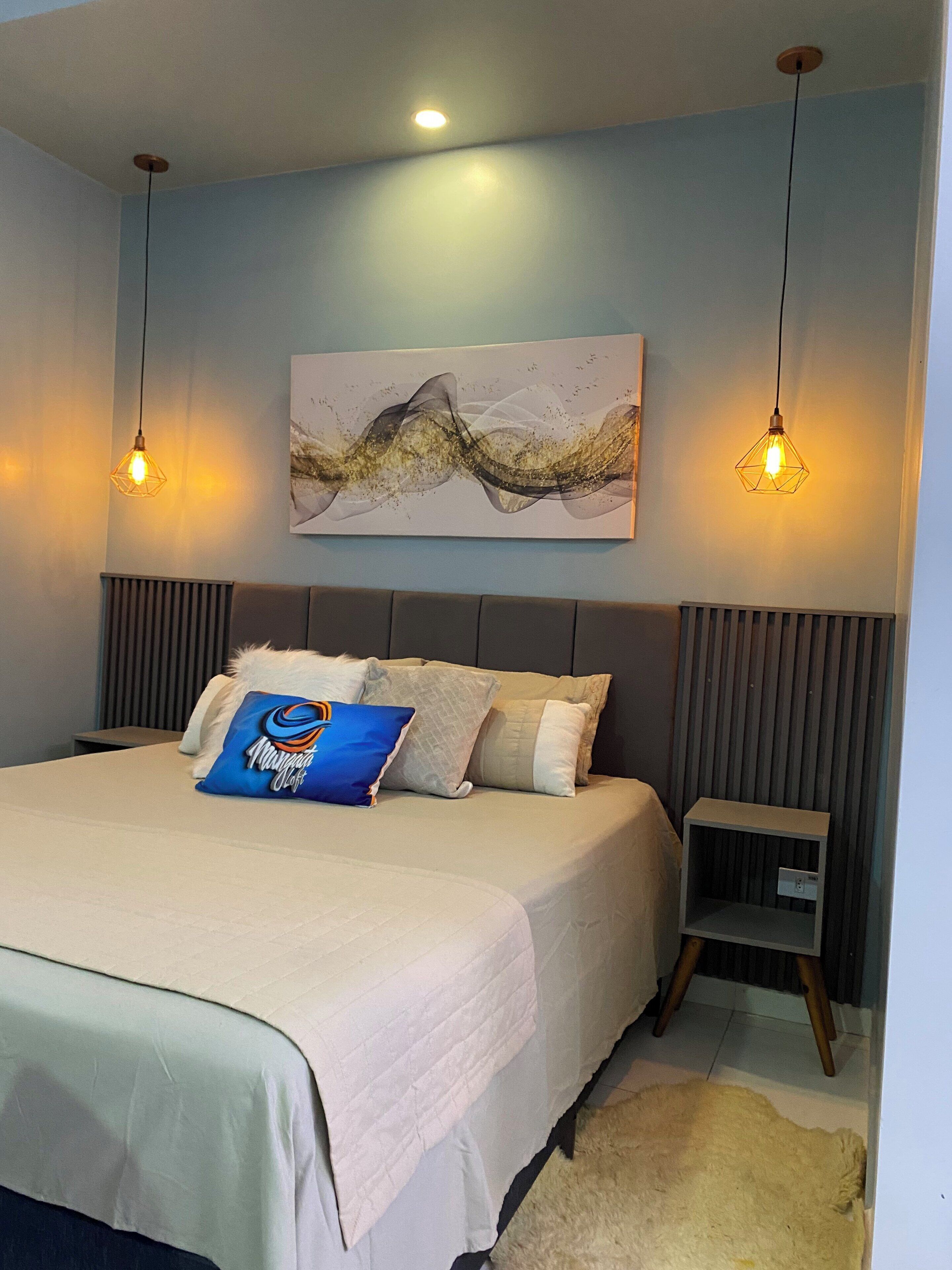 A modern bedroom with a bed, two pendant lights, a wall-mounted artwork, and a bedside table.