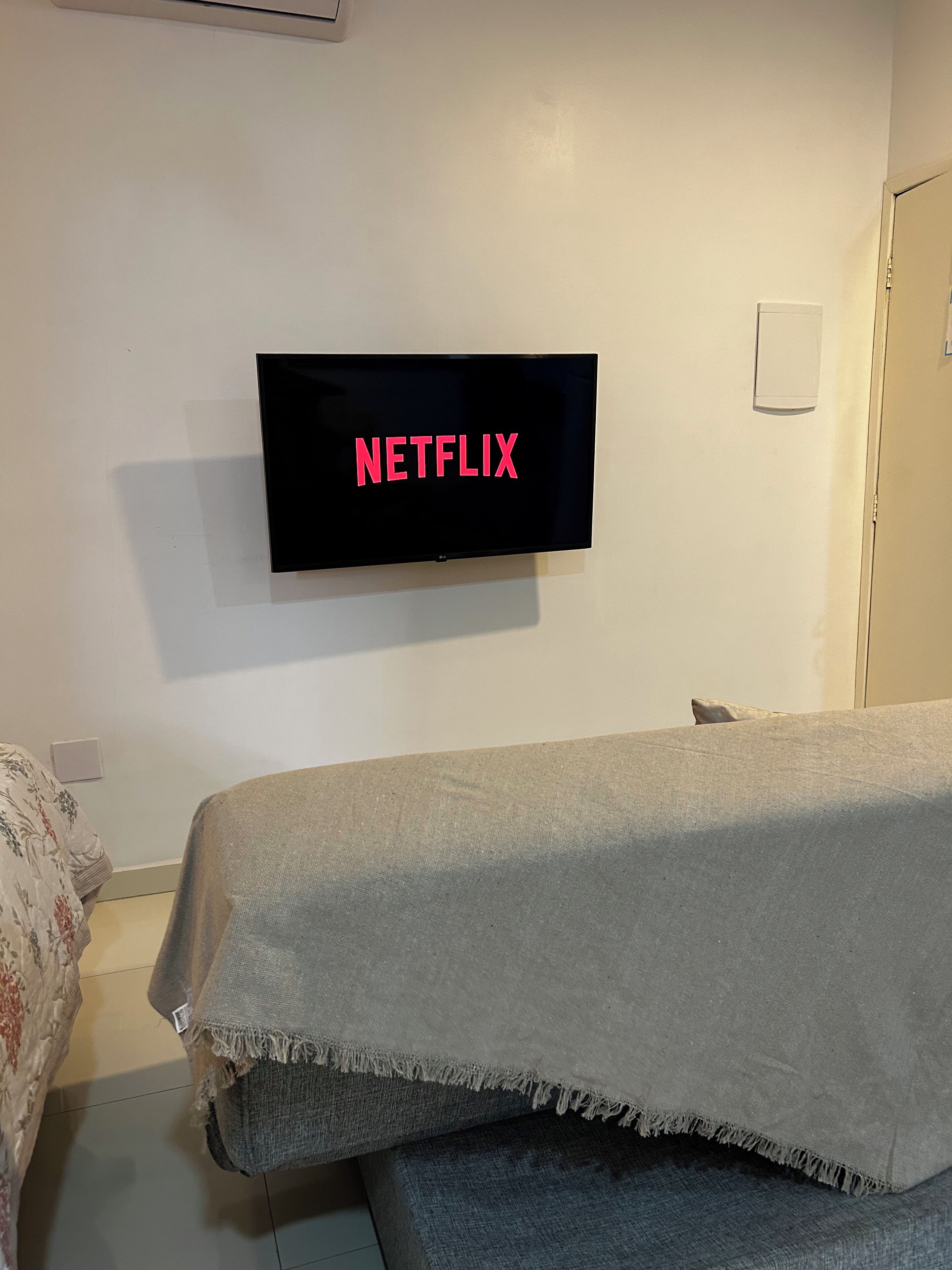 Een kamer met een bed, een aan de muur bevestigde televisie waarop het Netflix-logo te zien is, een deur en een lichtschakelaar.
