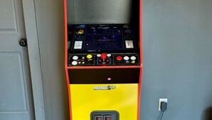Une borne d’arcade Pac-Man avec une base jaune et un dessus rouge, dotée d’un joystick et de boutons, placée devant un mur gris et sur un sol en bois.