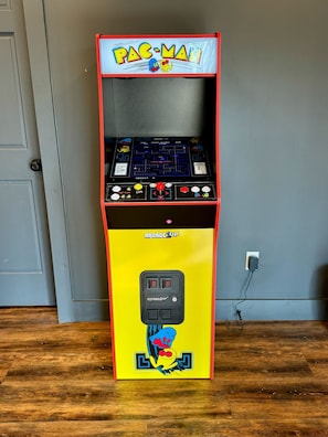 A Pac-Man arcade machine with a yellow base and red top, featuring a joystick and buttons, against a grey wall with a wooden floor. - The Roosevelt | Modern Cabin in the Trees (Logan)