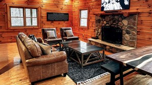A log cabin interior with a stone fireplace, leather sofa, and a television mounted on the wall. - The Roosevelt | Modern Cabin in the Trees (Logan)