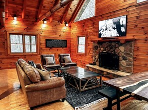 A log cabin interior with a stone fireplace, leather sofa, and a television mounted on the wall. - The Roosevelt | Modern Cabin in the Trees (Logan)