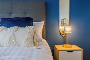 A neatly made bed with white and blue pillows, a bedside table with a lamp, and a mirror on the wall. - Toast Stays - The Sunroom House (Durham)