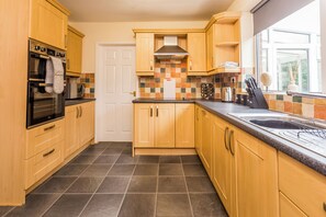 A modern kitchen with wooden cabinets, a stainless steel oven, a built-in microwave, and a large window providing natural light. - Toast Stays - The Sunroom House (Durham)