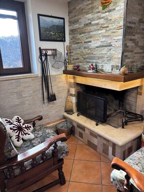 A cozy living room with a fireplace, a wooden bench, and a view of mountains through a window. - Discover the relaxation of the mountains in this detached villa surrounded by greenery. (Abruzzo)