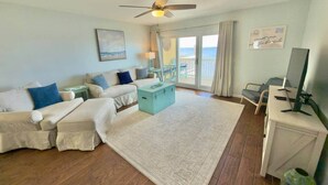 A spacious living room with a large window, a ceiling fan, a sofa set, a coffee table, and a desk with a computer. - Pelican's Perch: Kayaks, Views & More! (Escambia County)