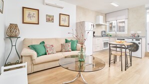 Un salon moderne avec une table basse en verre, un canapé beige agrémenté de coussins à motifs, un coin repas avec une chaise en métal et une cuisine équipée d’appareils électroménagers blancs.