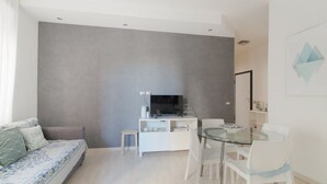A modern living room with a sofa, a glass dining table, and a TV on a white cabinet. - GuestHost - Cute Apartment Navigli Milano (Milan)