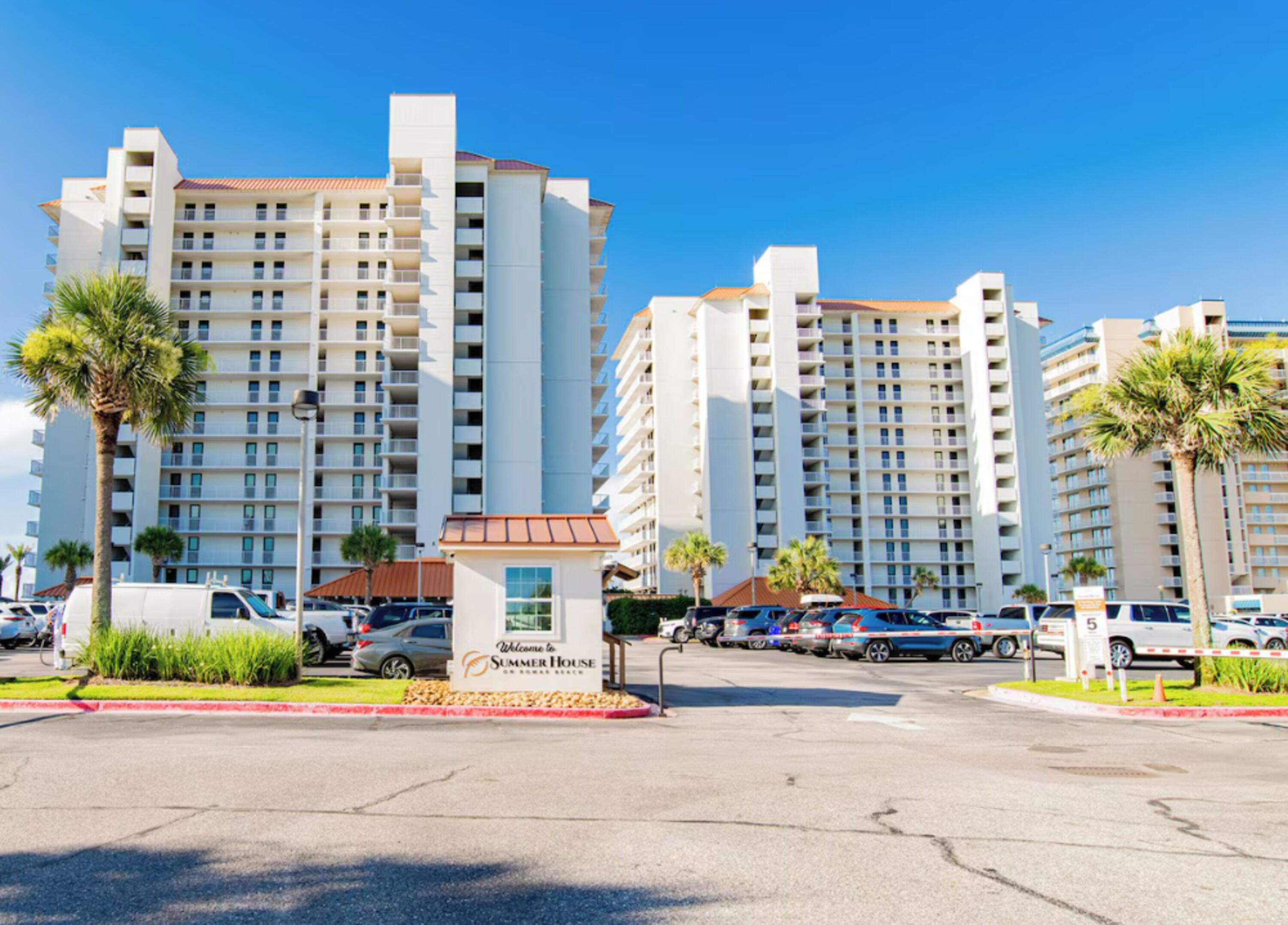 A multi-story hotel with a central entrance, parking area, and surrounding palm trees.