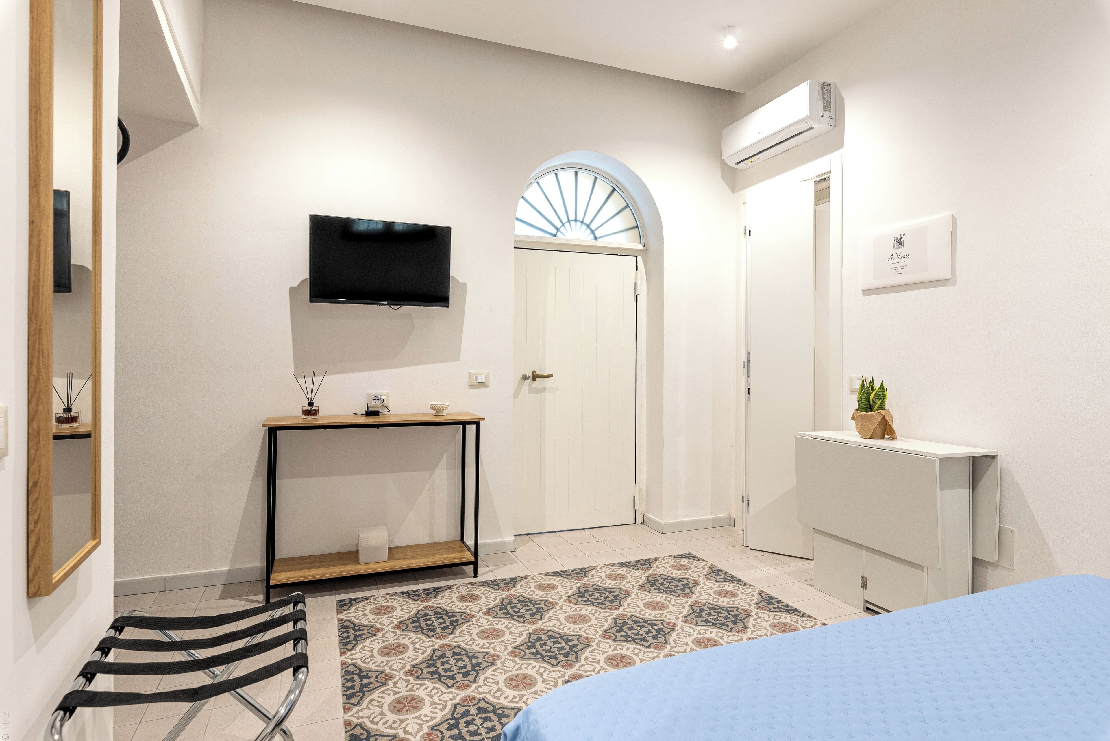 A modern bedroom with a bed, a TV mounted on the wall, a console table, a door with a fan, and a radiator.