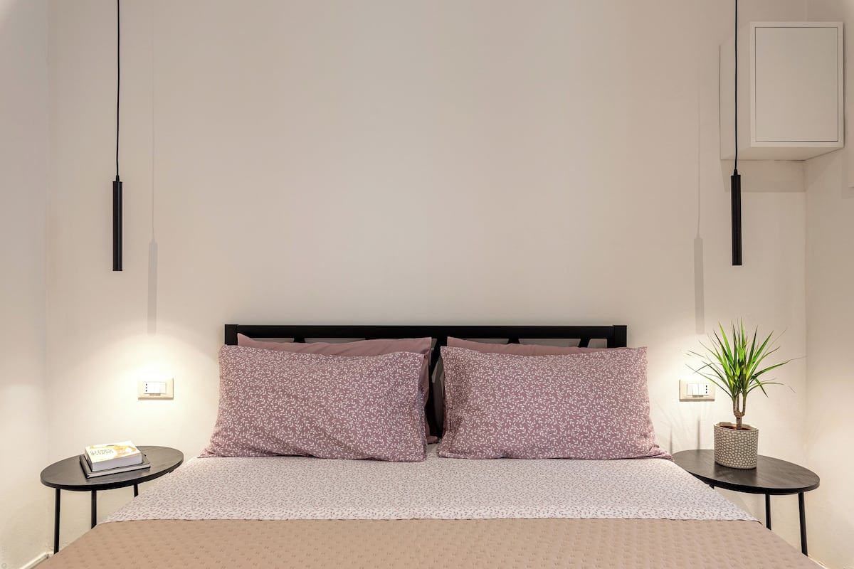 A neatly arranged bedroom with a bed, two bedside tables, a nightstand, a wall-mounted light, and a potted plant.