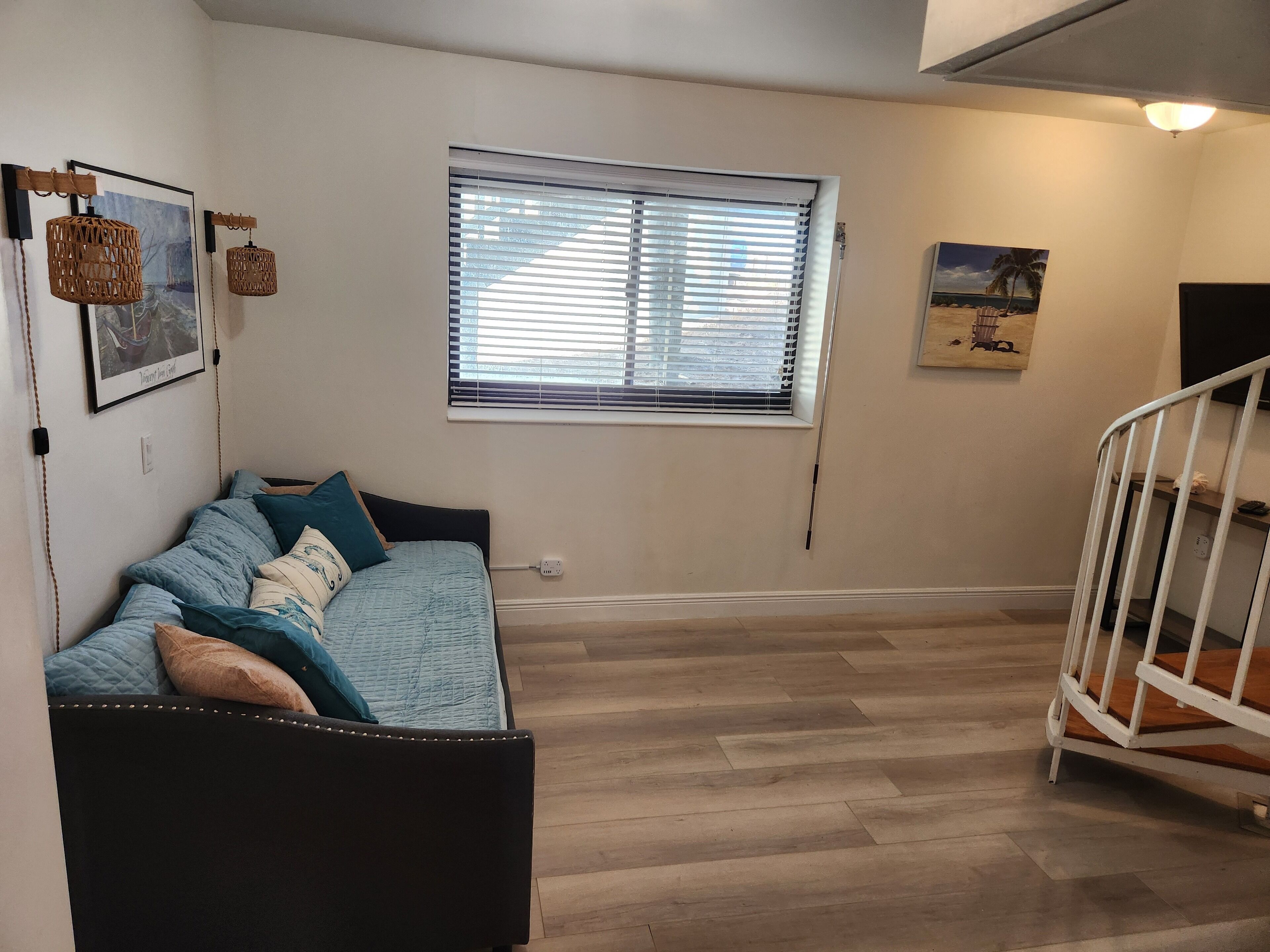 A cozy room with a bed, a window with blinds, a white railing, and a wall-mounted light fixture.