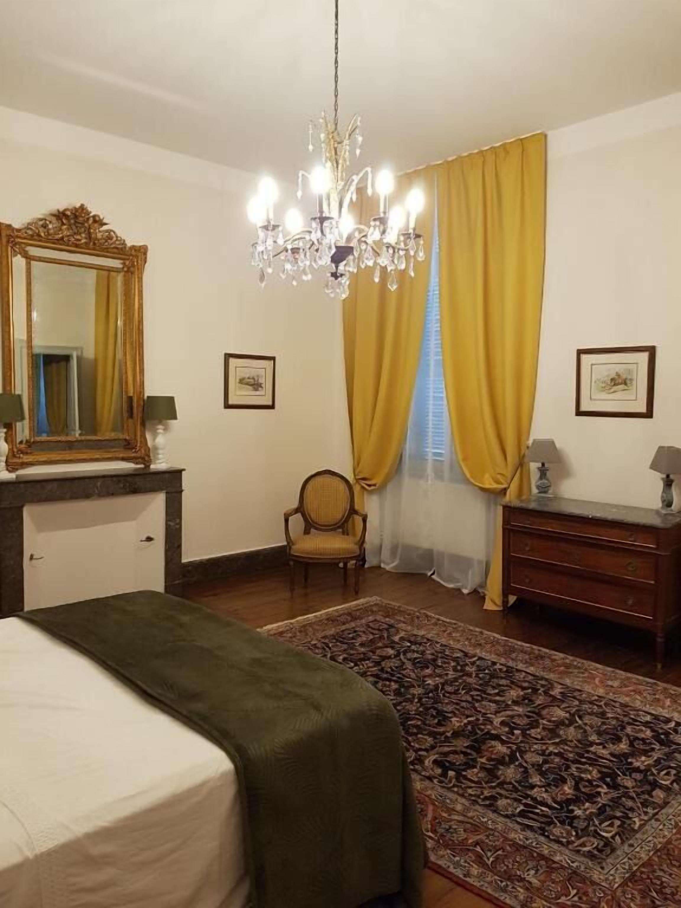 A bedroom with a bed, a dresser, a chair, a chandelier, and a large mirror.