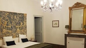 A bedroom with a bed, a chandelier, a large tapestry, a mirror, and a patterned rug. - Château St Justin (Saint-Justin)