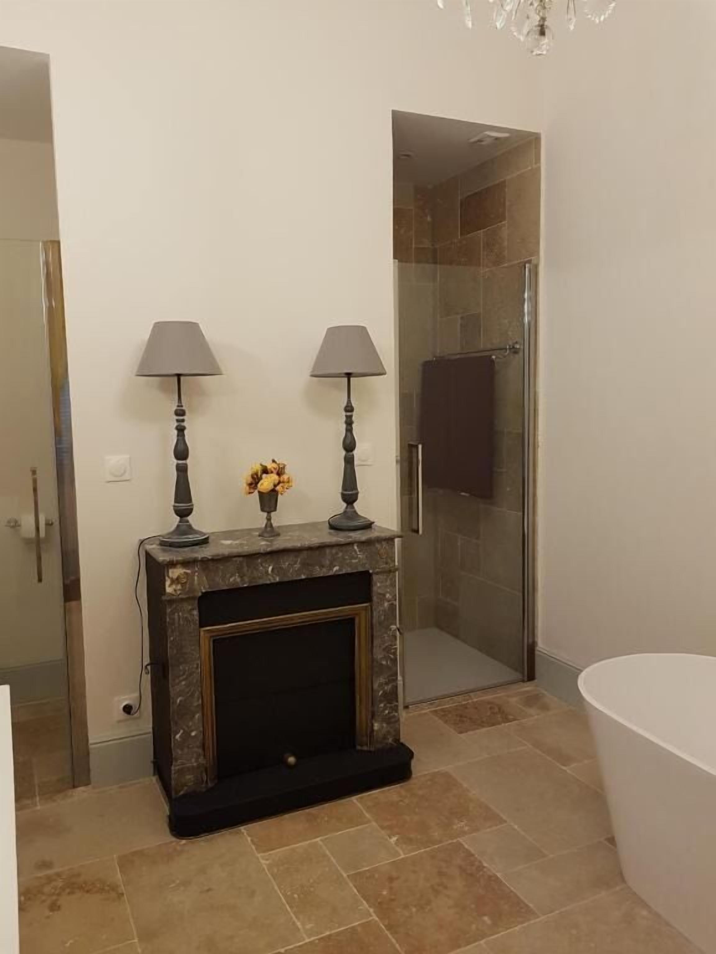 Un baño con cabina de ducha de vidrio, una bañera blanca y una chimenea de mármol con dos lámparas y un jarrón de flores.