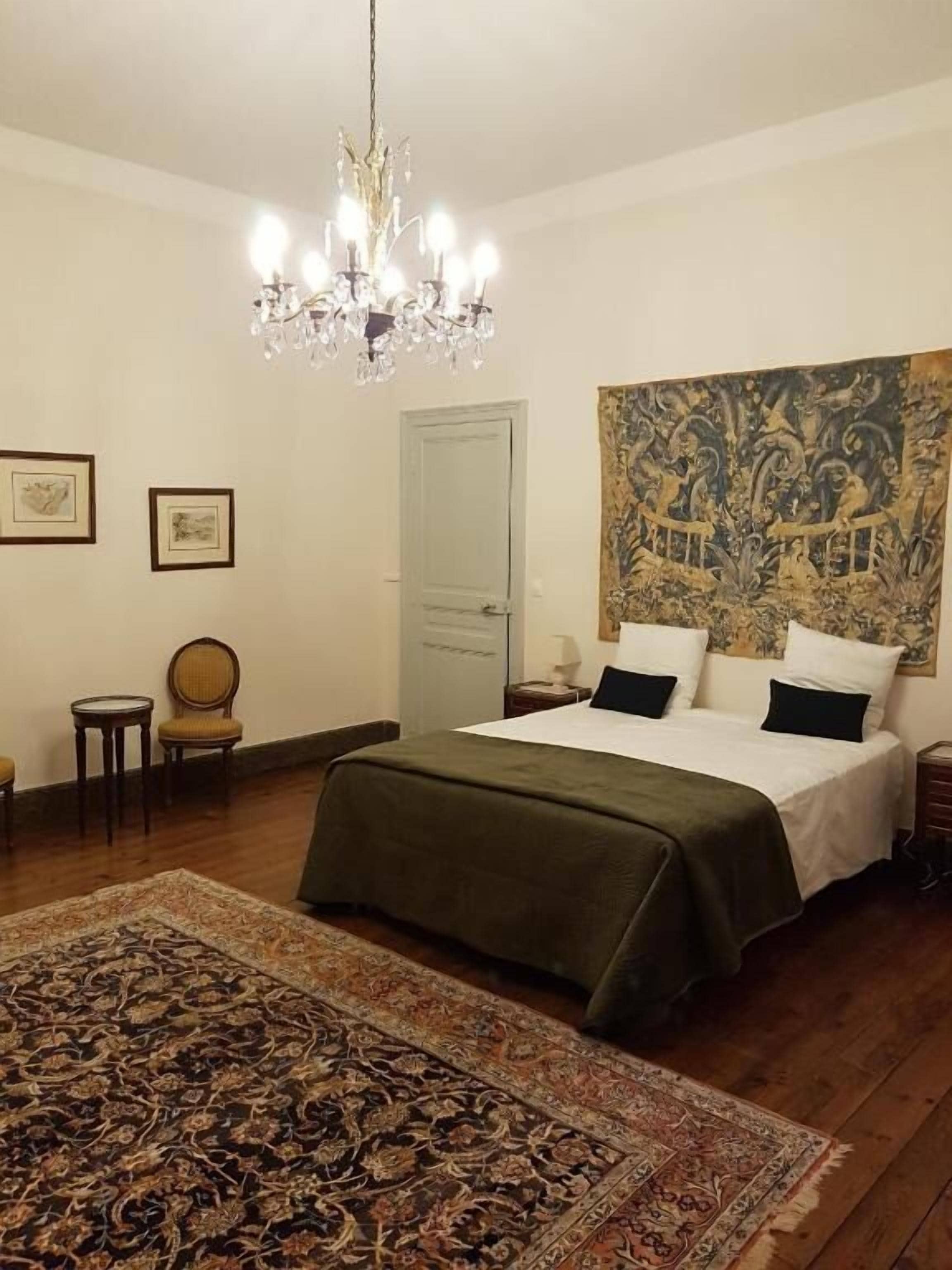 A bedroom with a large bed, a chandelier, a tapestry, and a patterned rug.
