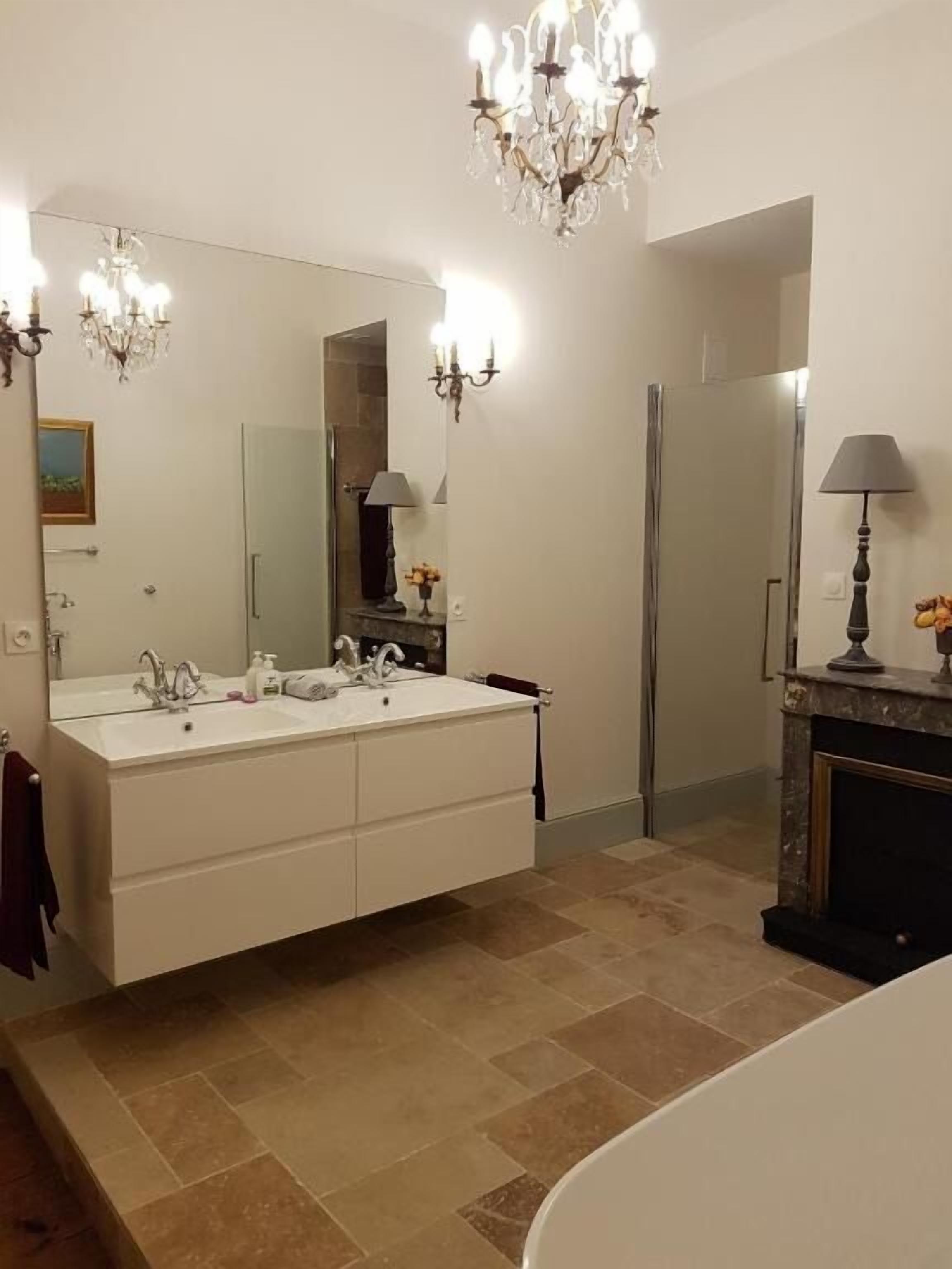 A bathroom with a double sink vanity, a large mirror, a chandelier, a fireplace, and a shower area.