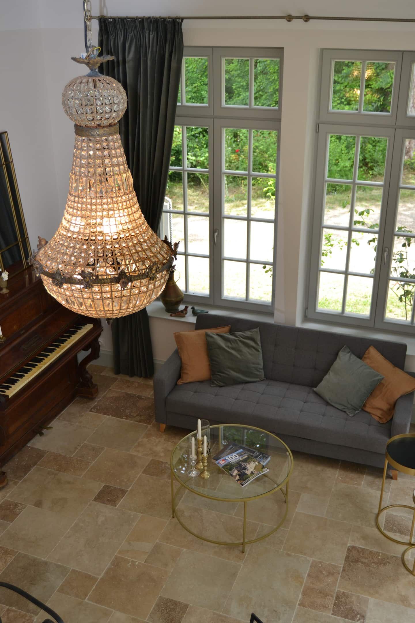 Un salón con un piano de cola, una araña, un sofá con cojines, una mesa de centro de cristal y amplias ventanas con vistas al verdor.