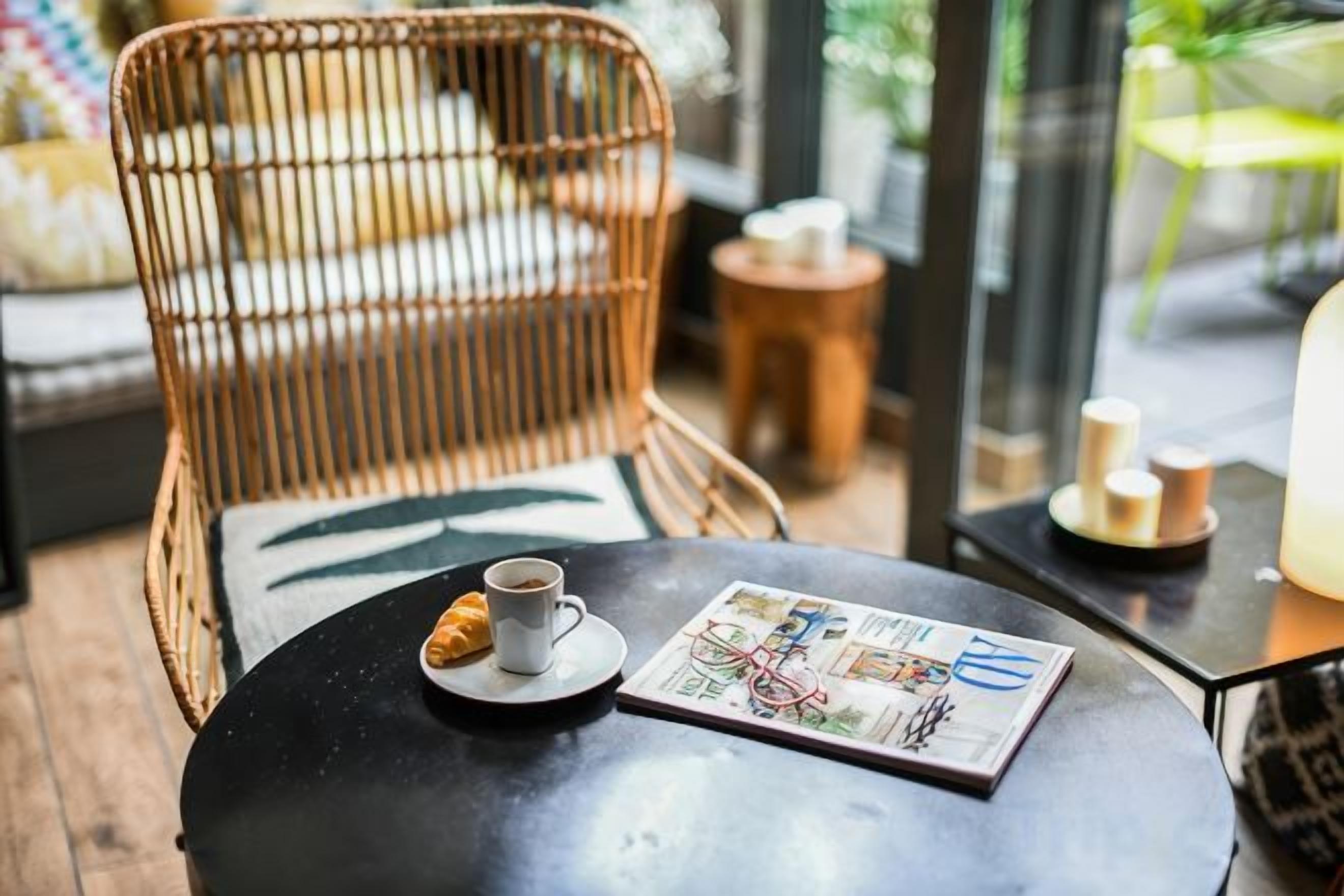 Una silla de mimbre con cojín, una mesa redonda con una taza y un menú, y una mesita auxiliar con velas.