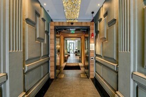 A hotel corridor with a central elevator, flanked by walls with paneling and a patterned ceiling. - Legend Hôtel Paris (Paris)