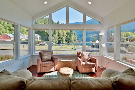 A sunlit room with large windows, two armchairs, a small ottoman, and a view of a lake and mountains.