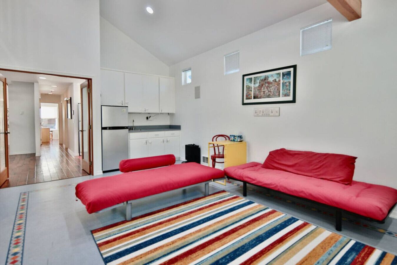 A compact room with a kitchenette, a sofa, a bench, and a colorful striped rug.