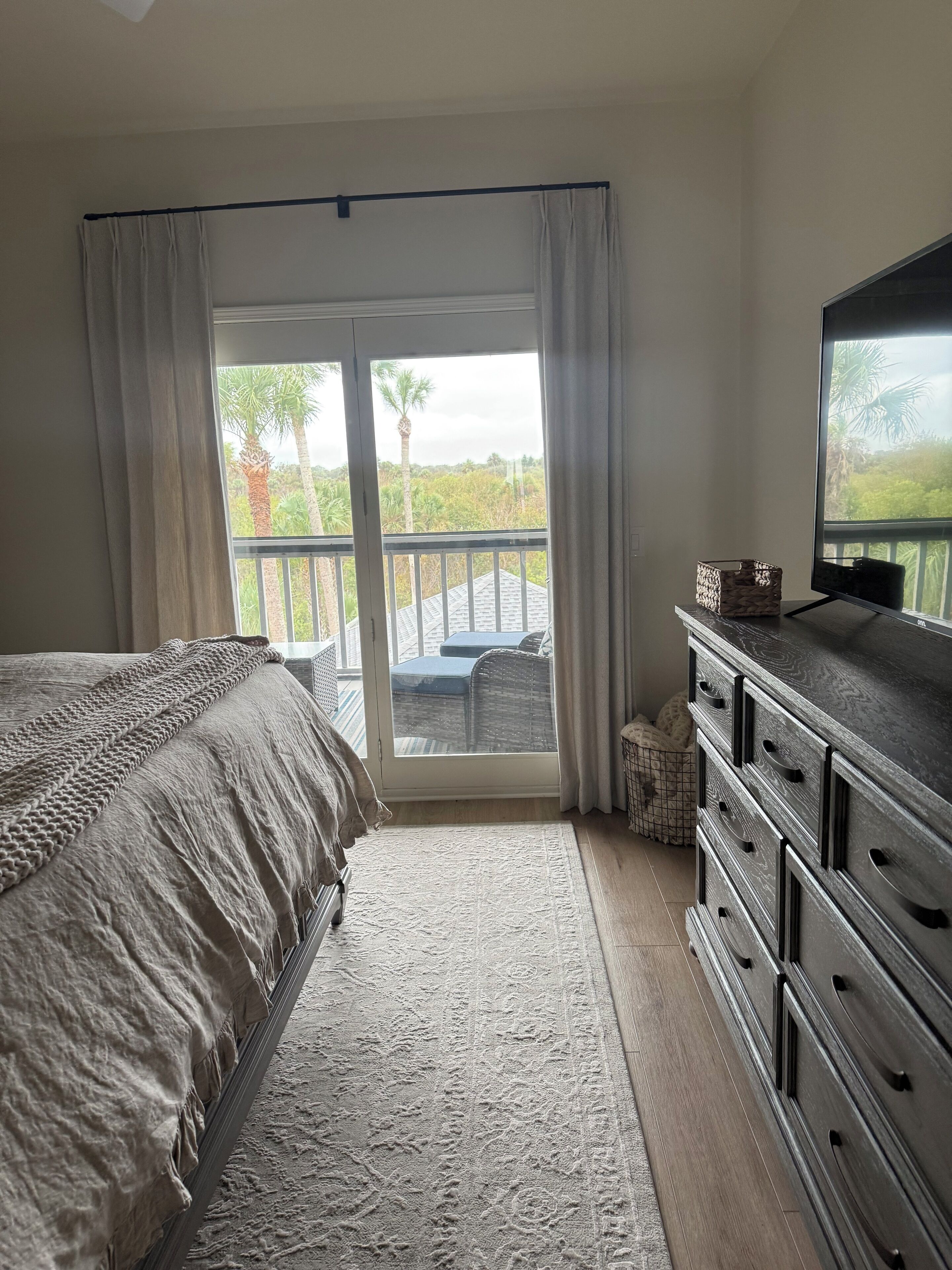 A bedroom with a bed, a dresser, a television, and a balcony with a view of greenery.