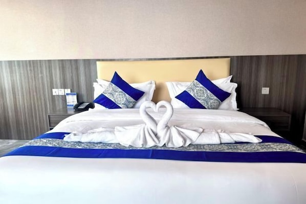 Un lit bien fait, agrémenté d’une décoration en forme de cœur réalisée avec une serviette, deux oreillers aux motifs bleus et blancs, et une table de chevet sur laquelle se trouvent un téléphone et une bouteille.