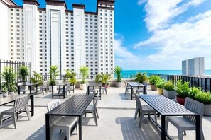 An outdoor patio with tables and chairs, potted plants, and a view of the ocean and tall buildings.