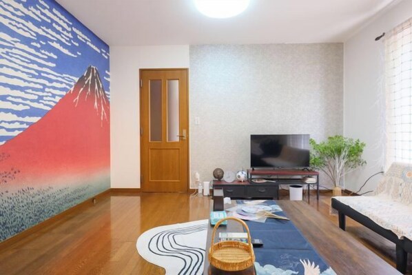 Une pièce avec un plancher en bois, une télévision, un canapé, un mur orné d’un tableau représentant une montagne et une porte.