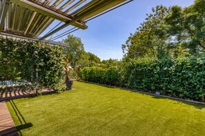 Une pelouse bien entretenue avec une terrasse en bois, entourée d’une végétation luxuriante et surmontée d’une structure de toit en métal.