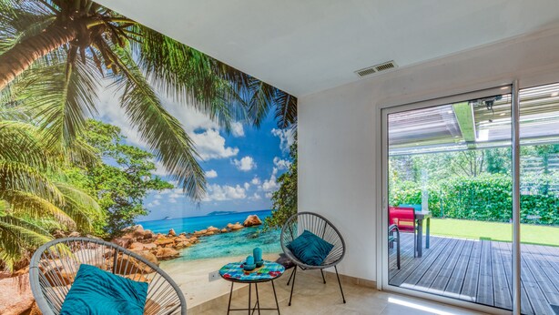 A room with a beach mural, two wicker chairs with cushions, a small table, and a sliding glass door leading to an outdoor deck.