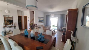 A spacious living area with a wooden dining table, white chairs, a sofa, and a large window. - Baleal Paradise Inn (Peniche)