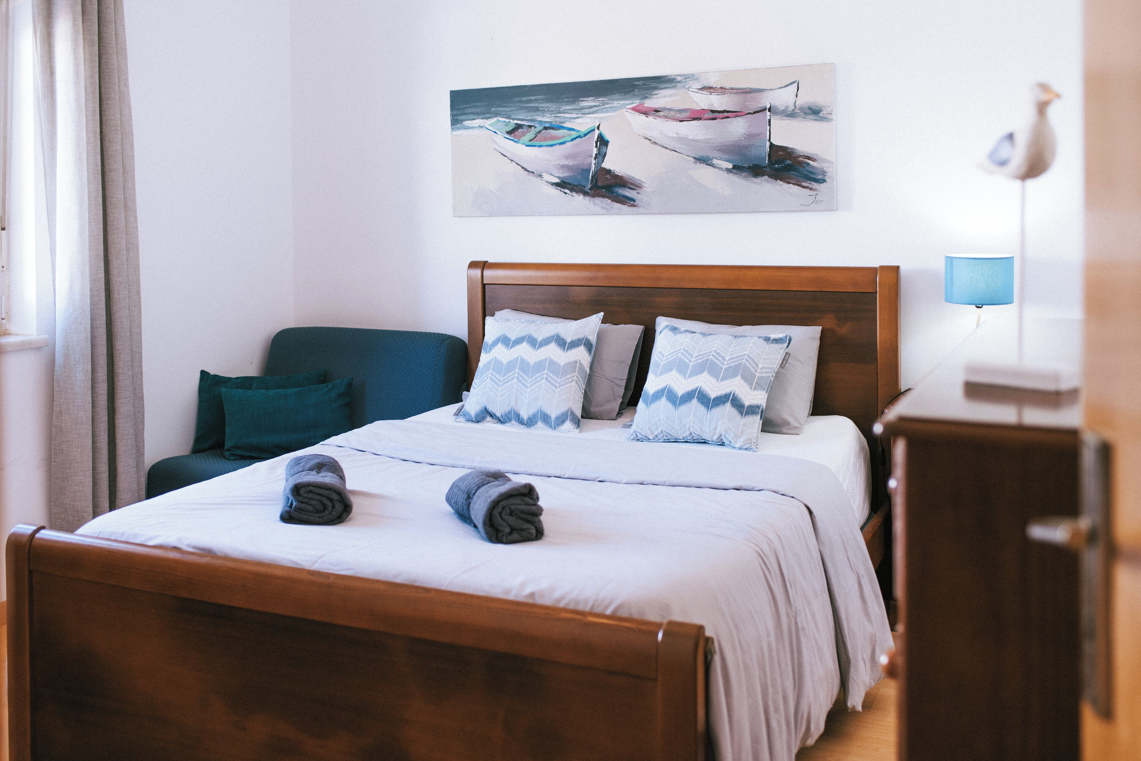 Ein ordentlich bezogenes Bett mit einem hölzernen Kopfteil, einer blauen Lampe und einem Gemälde von Booten an der Wand.