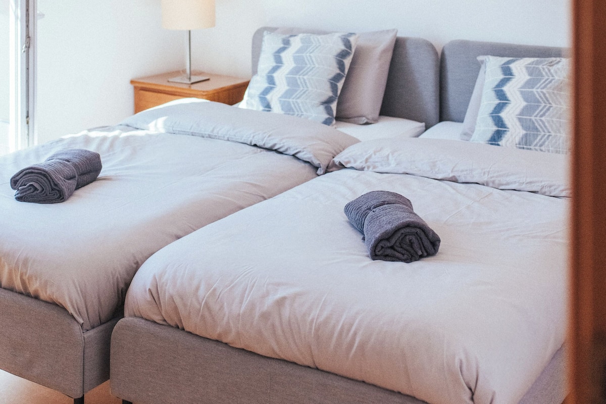 A bedroom with two beds, a nightstand, a lamp, and a beach-themed painting on the wall.