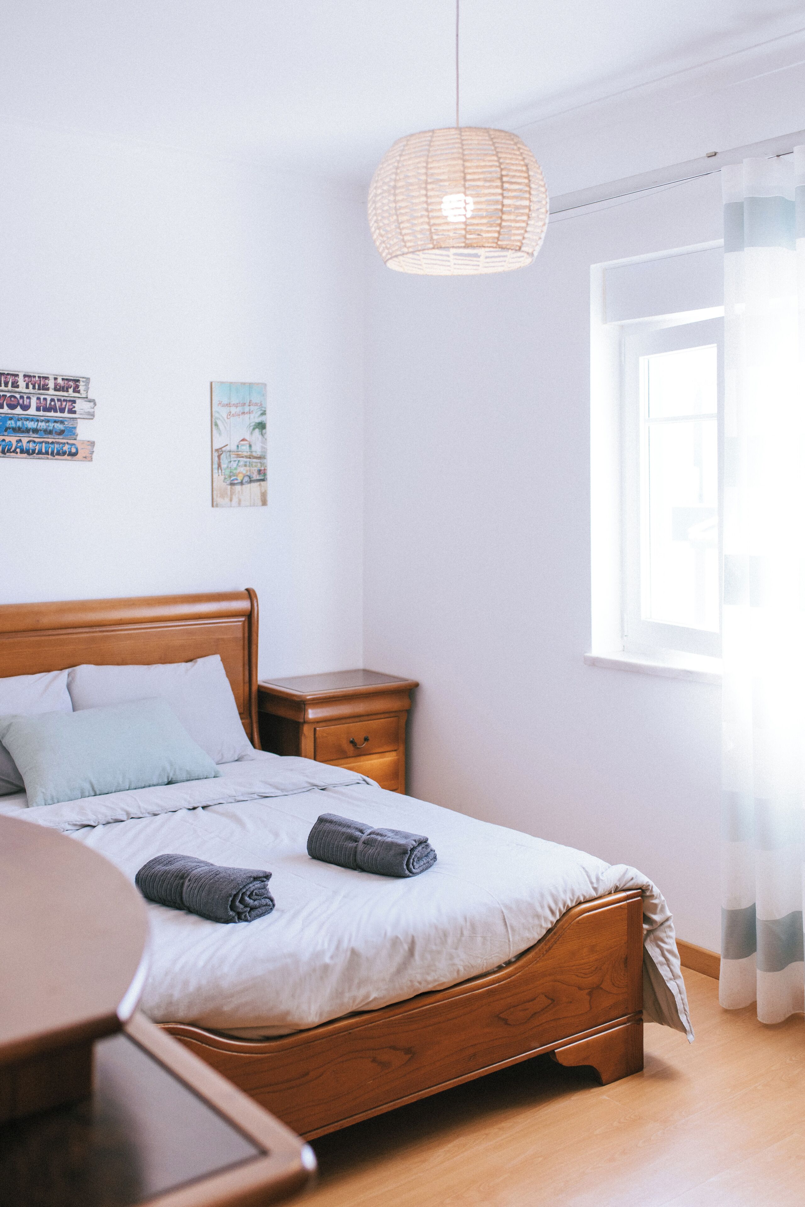 A neatly made bed with a wooden headboard, a bedside table, a hanging light fixture, and a window with curtains.