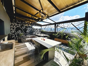 Un área de comedor al aire libre moderna con techo metálico geométrico, barra de concreto y pared con arte abstracto.