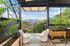 Un área de estar exterior moderna con sillas blancas y jardineras, con vistas a un paisaje urbano y montañas.