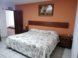 Un dormitorio con una cama, una mesita de noche, un cuadro y una puerta con la leyenda 'SALIDA DE EMERGENCIA'.
