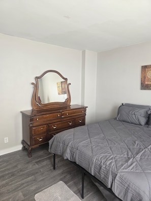 Une chambre avec une commode en bois, un lit avec une literie grise et un petit tapis sur le sol.