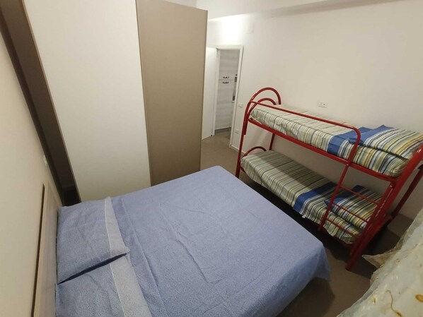A small, narrow room with a bunk bed and a single bed. There is a door leading to another room.
