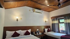 Une chambre d’hôtel avec deux lits, des poutres apparentes en bois au plafond, un climatiseur fixé au mur, des lampes de chevet, une fenêtre avec des rideaux et une petite table avec des articles de toilette.
