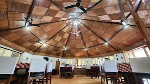 Une grande salle dotée d’un haut plafond en bois et de plusieurs ventilateurs de plafond. Des tables et des chaises sont disposées pour les repas ou les réunions.