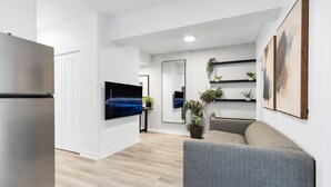 Een moderne woonkamer met een grijze bank, een flatscreen televisie aan de muur, een koelkast en een houten vloer.