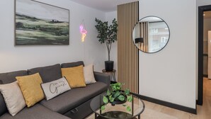 Egy modern nappali, melyben szürke színű kanapé, üveg asztallap, virágtartóban lévő növény, neonszínű flamingó alakú tábla, keretezett tájkép és egy kerek tükör található.