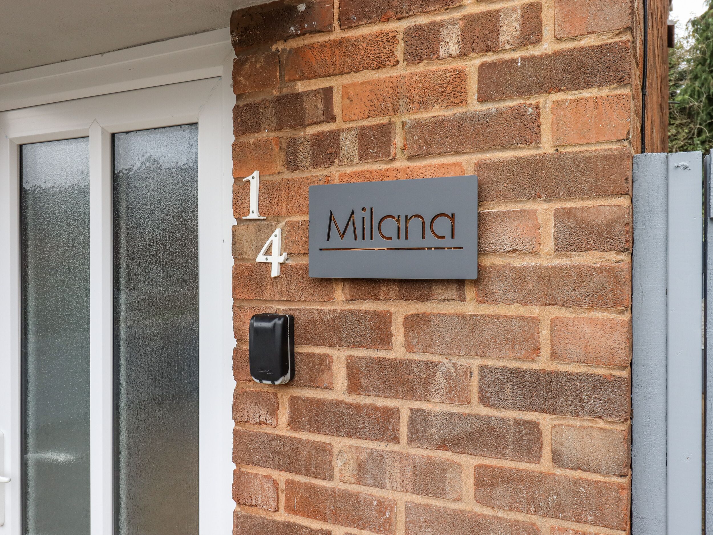 A brick wall with a house number plaque reading "14 Milana" and a black intercom unit.