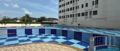 A swimming pool with blue tiles, a wooden deck, and a white building in the background.