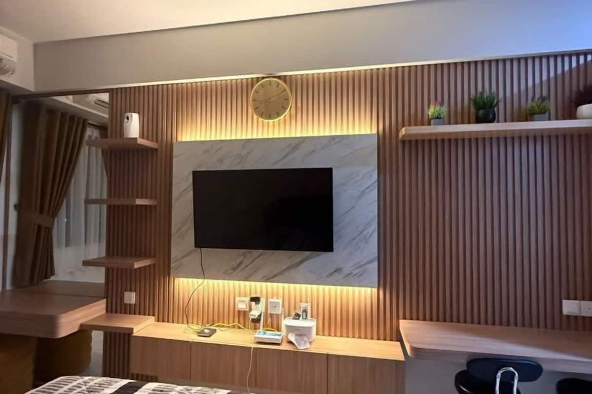 Una habitación de hotel moderna con un televisor de pantalla plana montado en una pared de paneles de madera, un escritorio de madera con una silla y un pequeño estante con plantas.